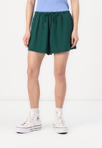 Shorts verdi in tessuto texturizzato, con vita elastica e cordino. Abbinati a sneakers alte bianche e una maglietta azzurro chiaro.