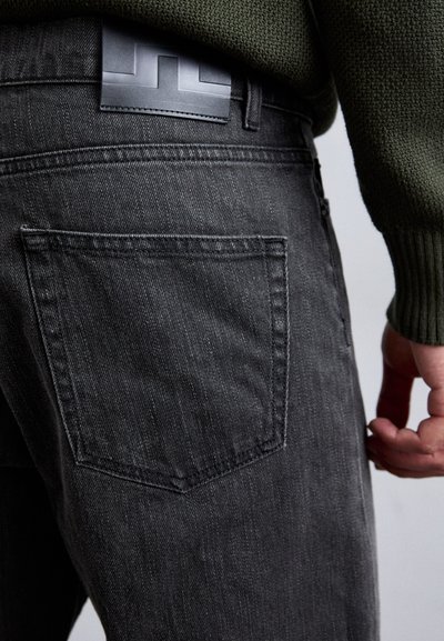 Dark gray denim jeans featuring a back pocket and a leather patch with a geometric design, paired with a textured green sweater.