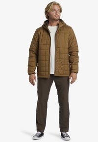 Brown quilted jacket with a hood, featuring a front zipper and patch pocket. Paired with dark pants and black-and-white sneakers.