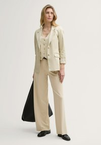 Crème-kleurig op maat gemaakt pak met een getailleerde blazer, bijpassend vest en wijde broekspijpen. Bevat knopen en een zwarte handtas.
