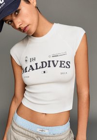 Hvid cropped t-shirt med navy tryk, der siger "MALDIVES," har korte ærmer. Sammen med grå sweatpants og en lys blå talje.