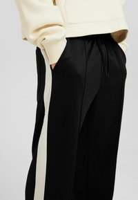 Svarta byxor med en vit sidostråk, tillverkade av slät tyg. Har resårmidja med dragsko och sidofickor.