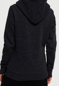 Persona vestida con un sudadera con capucha de color oscuro y moteado, vista desde atrás, de pie contra un fondo claro y liso.