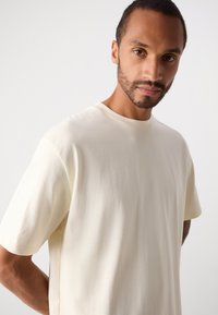 Uomo con capelli corti e barba curata che indossa una semplice t-shirt bianco sporco, in piedi su uno sfondo grigio chiaro.