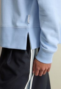 Lichtblauwe trui met geribde manchetten en zoom, zijsplitten en een labeldetail, gecombineerd met donkere korte broek met witte strepen.