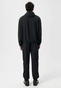Trening negru, caracterizat printr-o jachetă cu glugă, cu manșete și tiv elastic, asortat cu pantaloni care au buzunare tip cargo și manșete elastice la glezne.