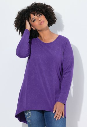 Kvinde med krøllet hår iført en lilla langærmet top og blå jeans, der rører ved sit hår, stående foran en ensfarvet lys baggrund.