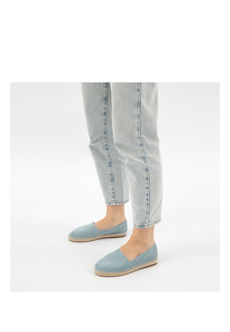 Person trägt hellblaue Jeans aus Denim und hellblaue Slip-On-Espadrille-Schuhe und steht auf einer weißen Fläche.