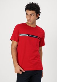 Camiseta roja de manga corta de algodón, con un panel gráfico que lleva el texto "TOMMY HILFIGER" y rayas horizontales en blanco y azul.