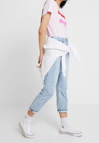 Rosa t-shirt med horisontella ränder i röd, vit och rosa; ljusblå kortdenimjeans; vit hoodie knuten runt midjan; vita sneakers.