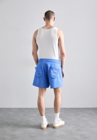 Pantalones cortos de algodón azul con dos bolsillos traseros, cinturilla elástica y detalle bordado. Llevado con una camiseta sin mangas blanca y zapatillas blancas.