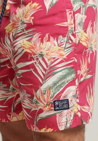 Bañador rojo con flores florales que presentan grandes flores tropicales en rosa y amarillo, con hojas verdes; incluye una etiqueta de logo "Superdry" en azul.