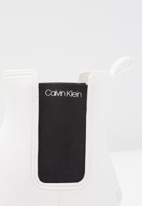 Botín blanco hasta el tobillo con un inserto elástico negro y detalles de costura, con un logo "Calvin Klein" en el elástico. Material suave y flexible.