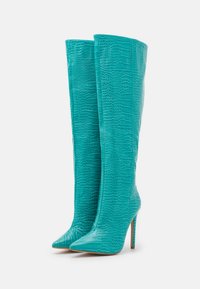 Botas altas de color turquesa con textura de cocodrilo, punta afilada y tacón de aguja alto. Superficie lisa con detalles de costura vertical.