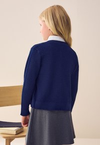 Marineblauwe gebreide cardigan met een gestructureerd patroon, cropped lengte en een gerafelde onderkant, gecombineerd met een grijze rok, gezien van achteren.
