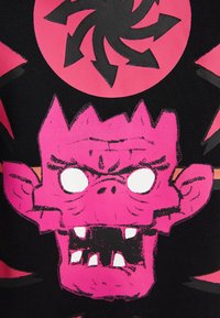 Ljust rosa, monstruös ansikte med tandställning, stora öron och ihåliga vita ögon på en svart bakgrund med en cirkulär design ovanför.