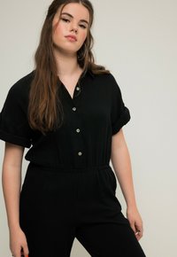 Černý krátký overal na krátké rukávy s předním zapínáním na knoflíky a elastickým pasem. Vyroben z texturované látky, s ohrnutými rukávy a plochými knoflíky.