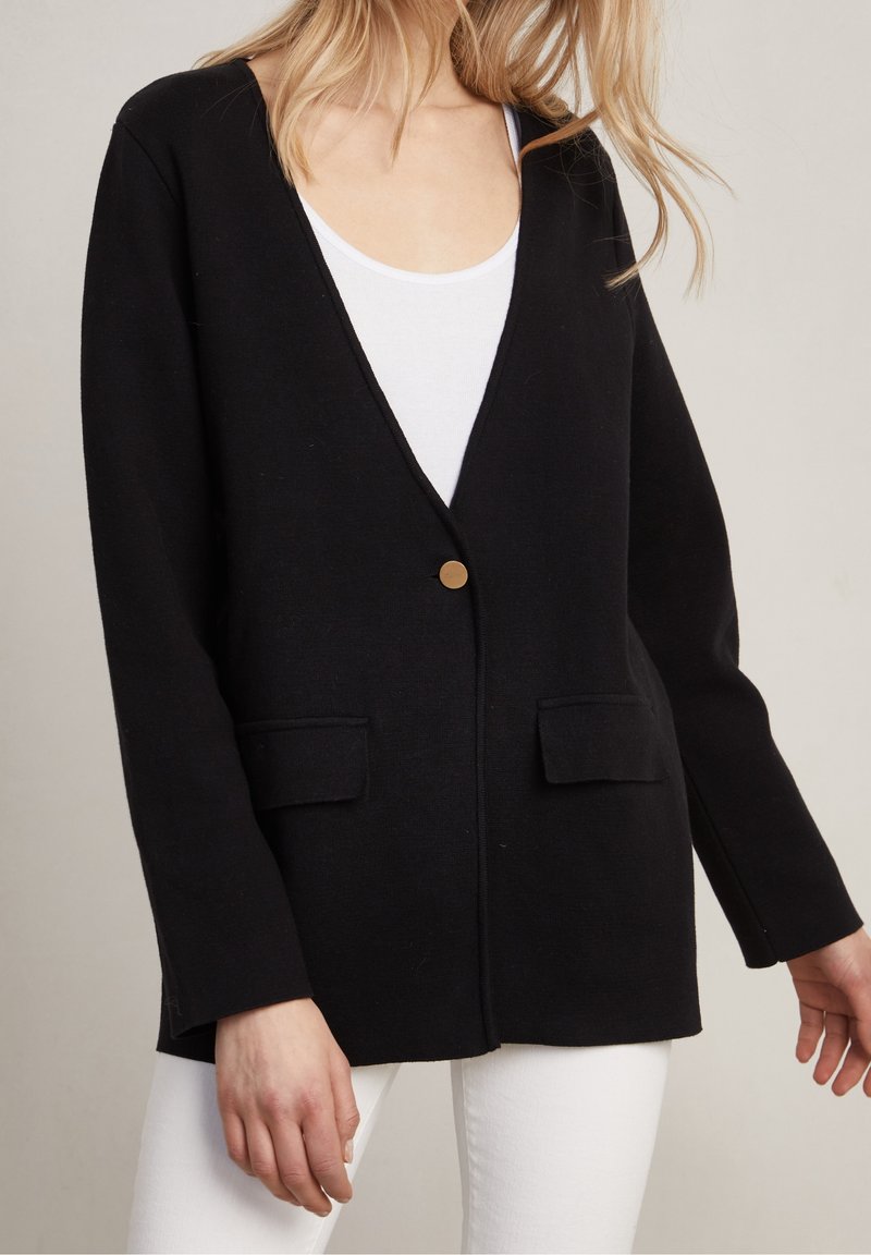 Femme portant un cardigan noir avec un seul bouton doré et des poches à rabat, par-dessus un haut blanc et un pantalon blanc.