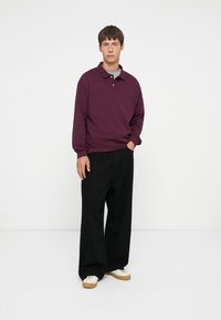 Dark purple long-sleeve polo shirt, relaxed fit, with a collar and three buttons, paired with wide black trousers and white sneakers.