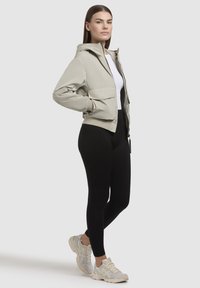 Light beige cropped jacket with a hood, two front pockets, streamlined design; paired with black leggings and beige sneakers.