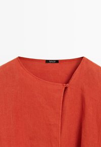 Orange Leinenbluse mit Rundhalsausschnitt, einem einzelnen Knopfverschluss nahe dem Kragen und einem sichtbaren Markenlabel am Halsausschnitt.