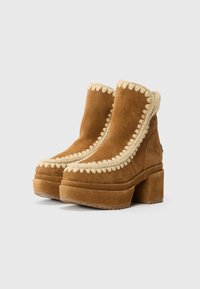 Stivaletti in suede marrone con suola platform e tacco a blocco, caratterizzati da eleganti rifiniture in crema a onde lungo il bordo superiore.