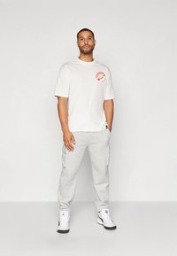 Balti trumpomis rankovėmis marškinėliai su raudonu piešiniu ant krūtinės, pilkos krovinių kelnės ir balti sportbačiai su juodais akcentais, stovintys priešais paprastą foną.