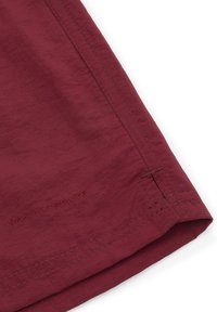 Burgundowa tkanina o teksturowanej powierzchni, z subtelnym przeszyciem i wytłoczonym logo blisko dołu. Dopasowany, prosty krój.