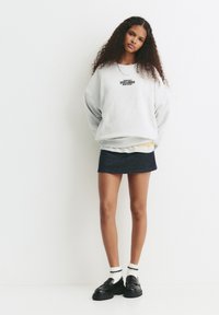 Felpa grigia oversize con grafica nera, indossata sopra una maglietta gialla, abbinata a una gonna di jeans scura e mocassini neri.