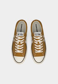 Ζεύγος χαμηλών παπουτσιών Converse All Star σε μουσταρδί κίτρινο χρώμα με λευκά κορδόνια και λαστιχένιες μύτες, από πάνω σε λευκό φόντο.