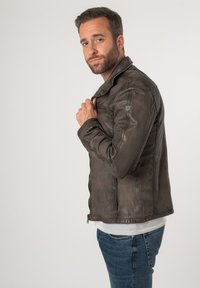 Giacca di pelle marrone con chiusura a zip, tasche laterali e vestibilità sartoriale, indossata sopra una camicia bianca e abbinata a jeans blu.
