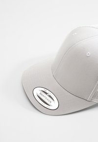 Hallikas snapback müts, millel on lame ääre, kangas esipaneel ja peenest võrgust tagapaneelid. Mütsil on ringikujuline silt, kus on kiri "THE CLASSICS SNAPBACK YUPOONG SINCE 1974."