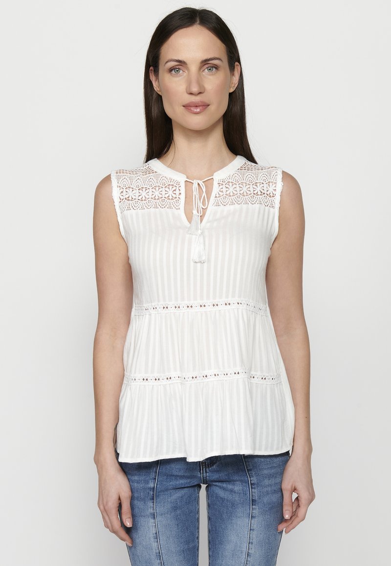 Top senza maniche bianco con inserti in pizzo e chiusura a fiocco, con righe verticali e volant a strati, abbinato a jeans blu.