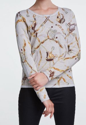 Osoba ubrana w jasnoszary sweter z długim rękawem z misternymi wzorami łańcuchów i kłódek w kolorze brązowym i złotym, w połączeniu z czarnymi spodniami, z założonymi skrzyżowanymi rękami.