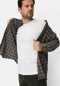 White crew-neck t-shirt under a dark plaid shirt with orange and blue accents, being pulled open. Model in dark green pants.