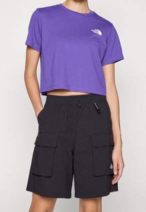 Person trägt ein lila kurzgeschnittenes T-Shirt und schwarze Cargo-Shorts mit Taschen, beide mit The North Face-Logo.