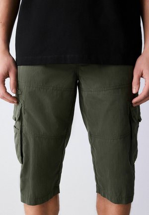 Man wearing olive green knee-length cargo shorts and a black t-shirt, standing with hands relaxed by sides.