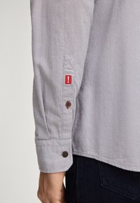 Camicia grigia a maniche lunghe con polsini abbottonati, caratterizzata da un'etichetta rossa laterale e bottoni marroni scuri. Il tessuto presenta una leggera texture e un motivo.