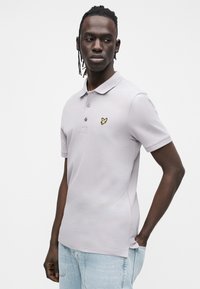 Polo grigio chiaro con scollo a collo, maniche corte e logo ricamato giallo sul petto a sinistra. Materiale in cotone, texture liscia.