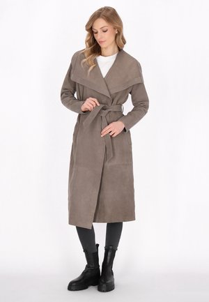 Vrouw die de riem vastmaakt van een lange taupekleurige trenchcoat, draagt zwarte leggings en zwarte enkellaarzen, staand tegen een witte achtergrond.