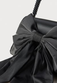 Bolso de mano de tela negra con un gran lazo decorativo y superficie texturizada, complementado con una correa de cuentas.