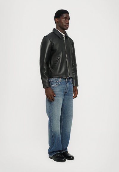 Chaqueta de cuero negra con cierre de cremallera, amplio cuello y bolsillos laterales, combinada con jeans de corte relajado en azul claro y zapatos negros.