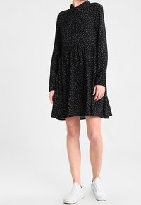 Robe noire à manches longues, col boutonné, taille froncée et motif à pois blancs. Complétée par des baskets blanches.