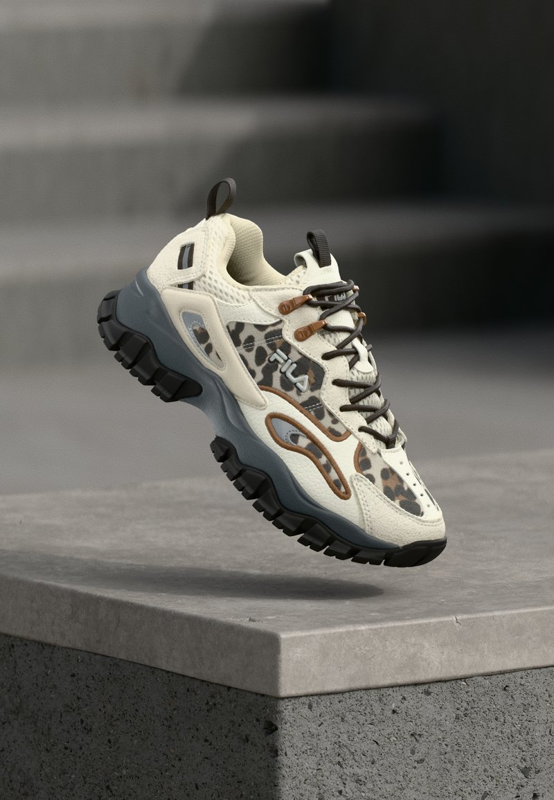 Baskets épaisses à imprimé léopard, accents beige et marron, semelle noire robuste, avec le logo FILA, en suspension au-dessus d’une surface en béton.