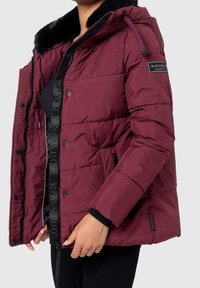 Vrouw in een donkerrode gewatteerde jas met een zwarte bontkraag, zwarte rits en een Navahoo-logo patch op de bovenarm.