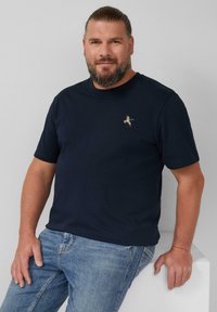 Man met een baard en samengebonden haar, gekleed in een marineblauw t-shirt met een klein geborduurd eendje en lichtblauwe spijkerbroek, leunt op een witte ondergrond.