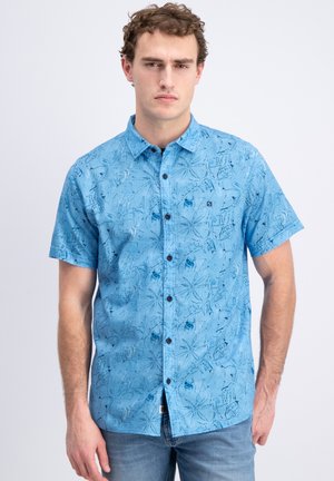 Ung mand iført en blå kortærmet skjorte med knapper og abstrakte blomster- og geometriske mønstre, stående foran en ensfarvet baggrund.
