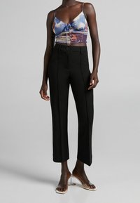 Kvinna som bär svarta högmidjade kortbyxor, en färgglad knyt-fram-topp och klara högklackade sandaler, står mot en grå bakgrund.