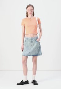 T-shirt cropped arancione chiaro, gonna in denim sbiadita con motivo in scrittura bianca, mocassini neri e calzini bianchi su uno sfondo semplice.