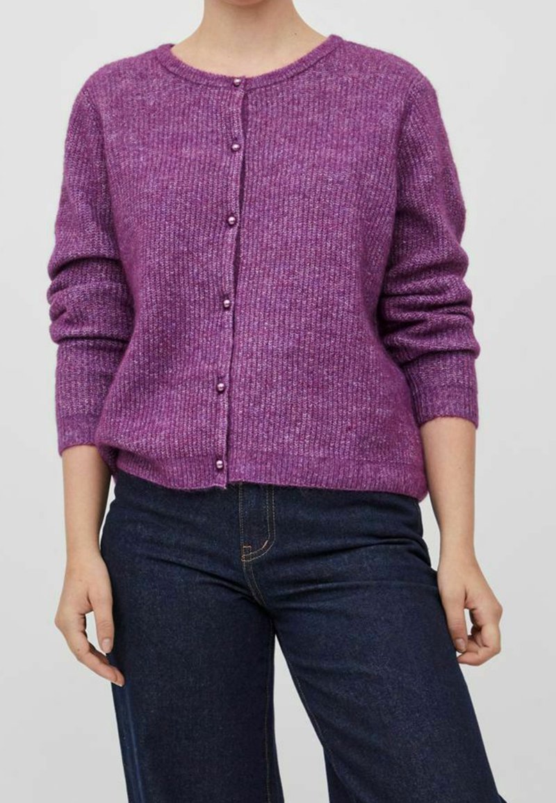 Femme portant un cardigan en maille violet avec des boutons ressemblant à des perles et un jean bleu foncé, debout devant un fond neutre.